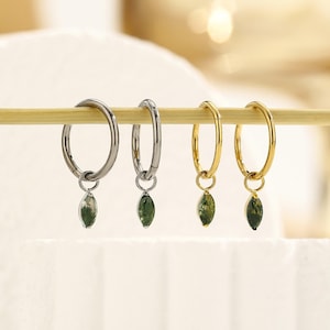 May include: Four hoop earrings, two silver and two gold, each with a green, leaf-shaped charm. The earrings are displayed on a wooden dowel against a soft, blurred background. The charms have a mossy appearance.