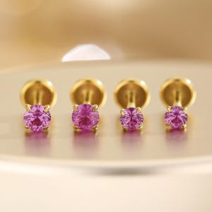 K&ouml;nnte beinhalten: Vier goldfarbene Ohrstecker mit runden, leuchtend pinkfarbenen Edelsteinen. Die Ohrringe sind in einer Reihe angeordnet und zeigen die lebendige Farbe der Edelsteine und die polierte Goldfassung. Die Ohrringe sind klein und zart.