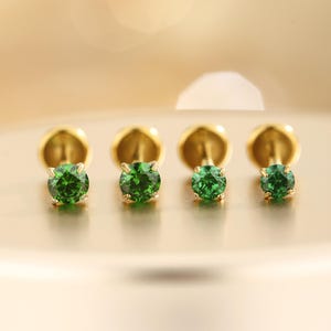 May include: Four gold-colored stud earrings with round, vibrant green gemstones. The earrings are set against a soft, golden background, highlighting the jewelry's sparkle and design. The studs are small and delicate, perfect for everyday wear.