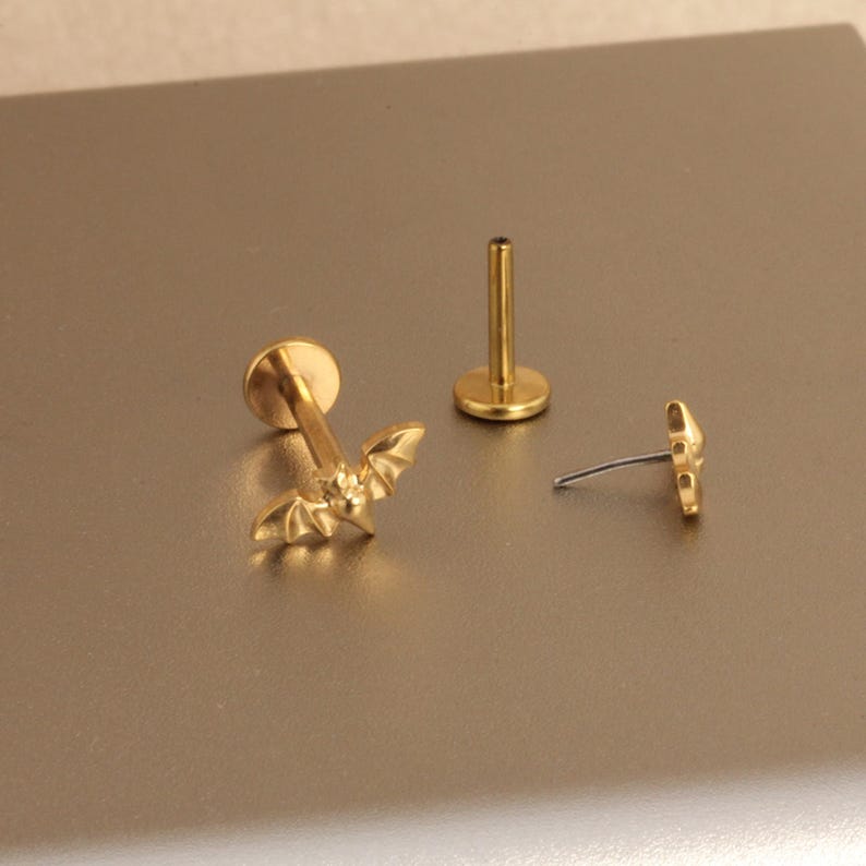 May include: Gold-colored lip piercing jewelry with a bat design. The jewelry includes a bat-shaped stud, a flat-backed stud, and a post. The bat has detailed wing features. The jewelry is displayed on a neutral surface.