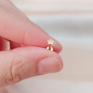 Op de afbeelding: Gouden ster oorsteker met een kleine, platte ster bedel. De oorsteker wordt in een hand gehouden.