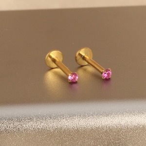 Może przedstawiać: Dwa złote kolczyki do piercingu warg. Każdy kolczyk ma mały, okrągły, r&oacute;żowy kamień szlachetny na końcu prostego pręcika. Kolczyki mają płaskie, okrągłe oparcie.