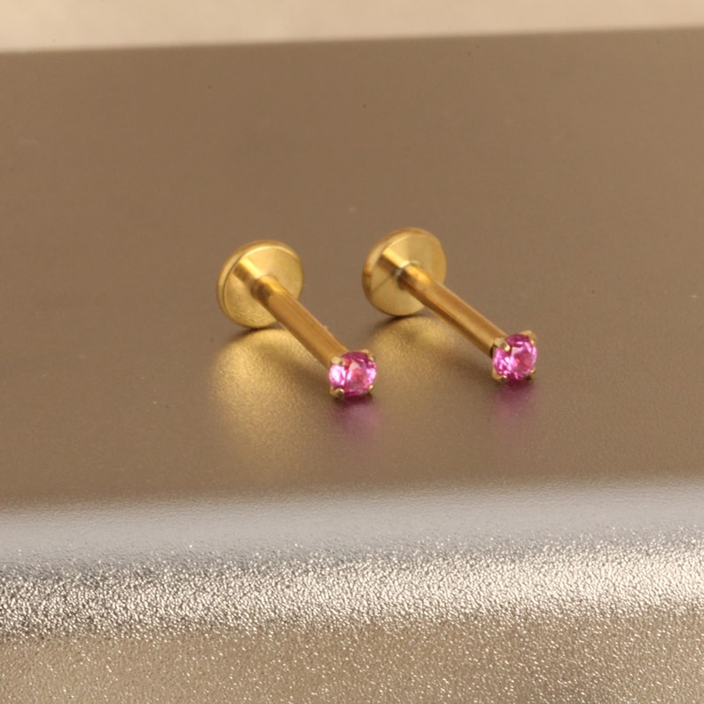 Może przedstawiać: Dwa złote kolczyki do piercingu warg. Każdy kolczyk ma mały, okrągły, r&oacute;żowy kamień szlachetny na końcu prostego pręcika. Kolczyki mają płaskie, okrągłe oparcie.