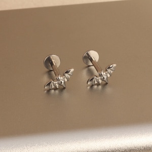 May include: Two silver-colored labret earrings. Each earring features a small bat design with outstretched wings. The earrings have a flat, round backing and a straight post for piercing. The bat's wings are detailed with etched lines.
