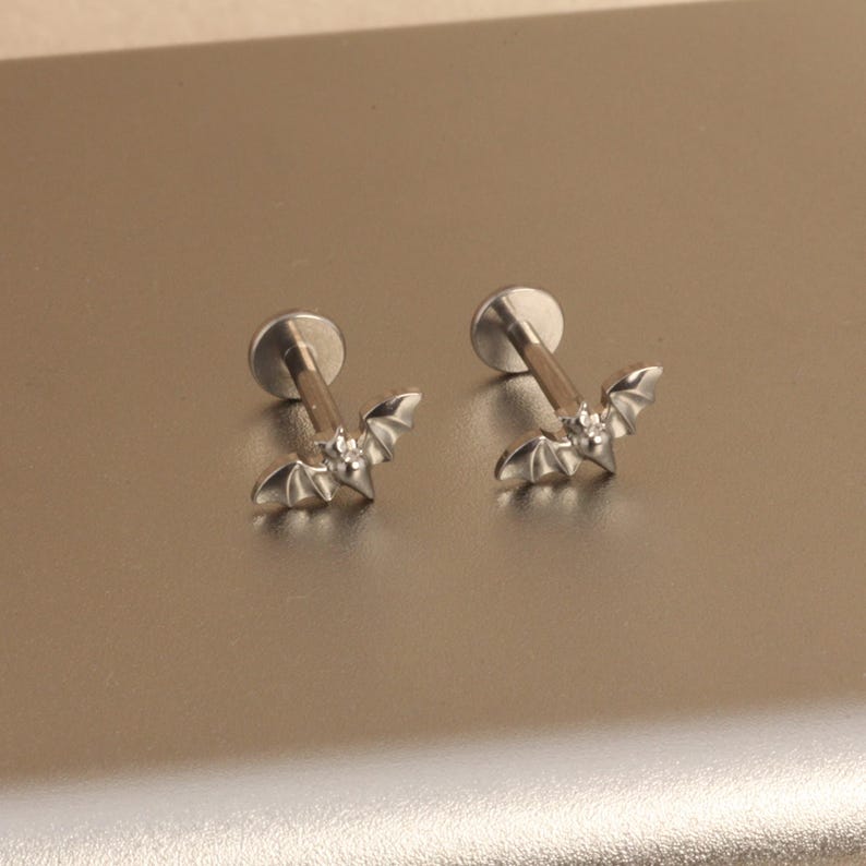 May include: Two silver-colored labret earrings. Each earring features a small bat design with outstretched wings. The earrings have a flat, round backing and a straight post for piercing. The bat's wings are detailed with etched lines.