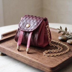 May include: A small, burgundy leather purse with a textured flap and a gold chain strap. The purse has pink accents and gold-toned hardware. It sits on a wooden tray with brass feet, showcasing a handcrafted design.