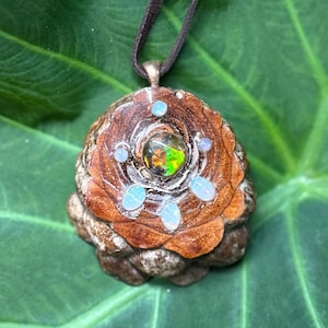 May include: A pendant crafted from a brown pinecone scale featuring a central round iridescent stone with green and orange flashes, surrounded by five small oval white opalescent cabochons, suspended from a dark brown cord.