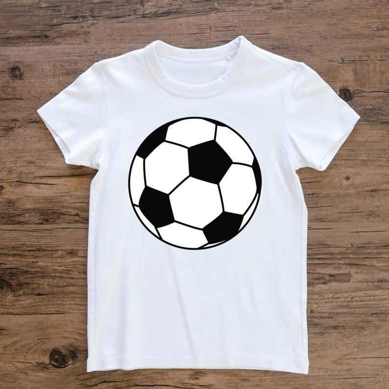 May include: White t-shirt with a black and white football design. The football graphic is centrally placed and features black pentagons and white hexagons, outlined in black. The shirt has short sleeves and a crew neck.