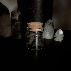 May include: A small glass jar with a cork lid filled with black crystals. The jar is sitting on a black surface with other clear crystals.
