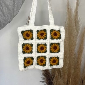Peut inclure: Sac fourre-tout en crochet blanc avec des motifs de tournesols. Le sac a une forme carrée et deux longues anses.