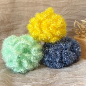 Può includere: Tre spugne rotonde e soffici nei toni del giallo, verde e blu. Le spugne hanno un aspetto strutturato e sfocato, con un filo d'argento intrecciato. Sono disposte su un tessuto morbido dai toni neutri.