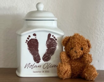 Puede incluir: Jarrón de cerámica blanca con tapa, con dos huellas de manos marrones y el texto "Nolan Oliver, 24 de septiembre de 2022". Un oso de peluche marrón se sienta junto al jarrón.