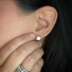 May include: A close-up of a person's ear with a small, round, diamond stud earring. The earring is set in a gold setting and has a halo of smaller diamonds around the center stone.