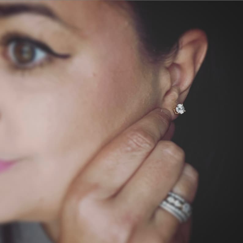 May include: A close-up of a woman's ear with a small, round, clear diamond stud earring. The earring is set in a gold setting.
