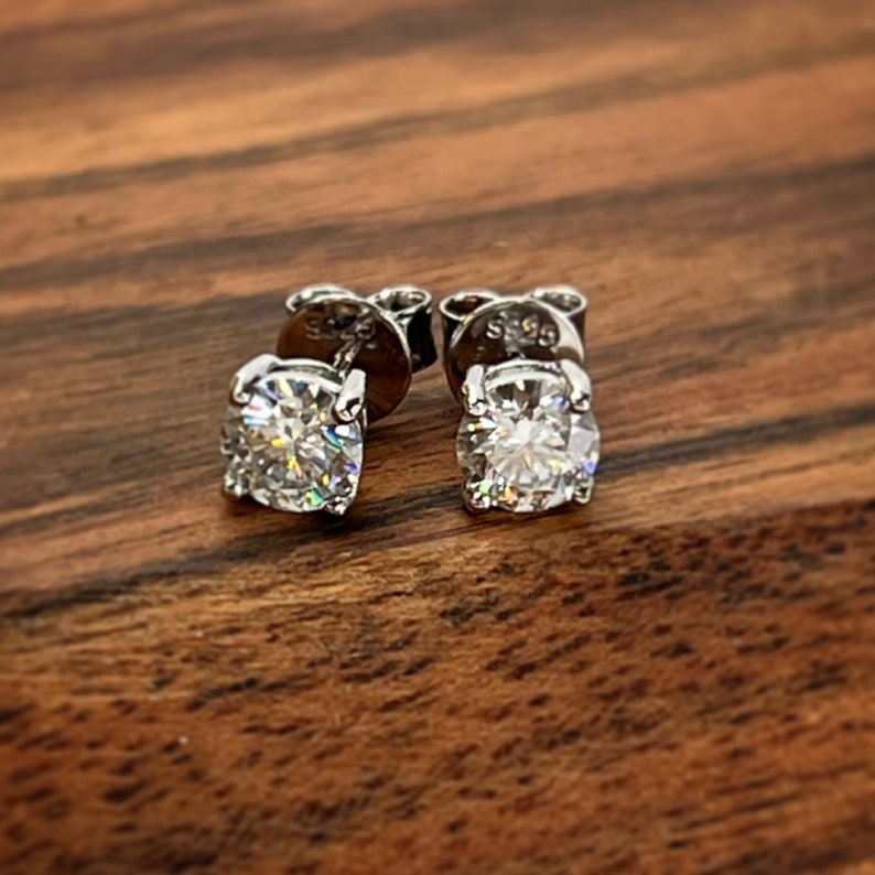 May include: A pair of silver stud earrings with clear round gemstones. The earrings are on a wooden surface.