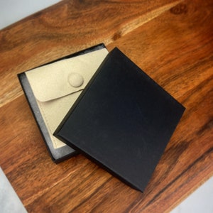 May include: A black jewelry box with a beige velvet interior and a button closure. The box is open and the interior is visible.