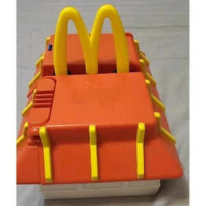 May include: A vintage McDonald's Happy Meal toy in the shape of a restaurant. The toy is primarily orange with yellow accents and the iconic golden arches logo. The base is white.