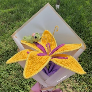 Puede incluir: Una flor de lirio amarilla de crochet con detalles morados, en un ramo de papel blanco, con una pequeña rana verde de crochet en la parte superior. El ramo está atado con una cinta rosa y tiene el texto "@crochetmerry" en la parte inferior.