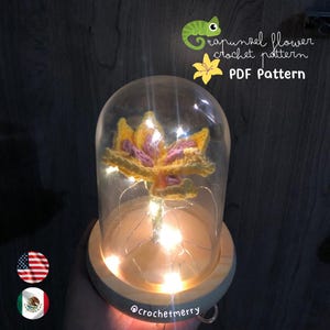 Peut inclure: Une fleur en crochet jaune et rose avec une tige à l'intérieur d'un dôme en verre sur une base en bois avec des lumières féériques. Le texte "Rapunzel flower crochet pattern PDF Pattern" est au-dessus du dôme. Le texte "@crochetmerry" est en dessous du dôme.