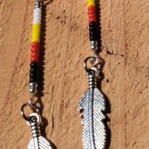 May include: Silver feather earrings with beaded accents in red, yellow, white, and black. The earrings have a hook closure and are displayed against a wooden background. These earrings are a unique accessory.
