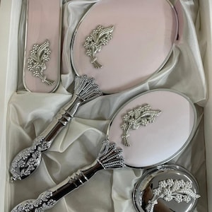 Puede incluir: Un juego de tocador con un espejo de mano rosa, un espejo compacto y un cepillo. Cada pieza tiene un diseño floral plateado y un mango plateado. El juego se presenta en una caja blanca con forro de satén.