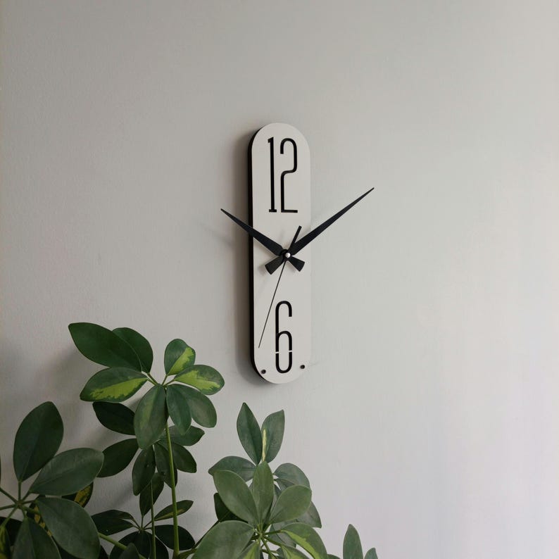 May include: A modern, minimalist wall clock with a white oval face and black hands. The clock features the numbers 12 and 6 in a sleek, black font. The clock is mounted on a light gray wall, with green leafy plants in the foreground.
