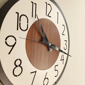 Puede incluir: Un reloj de pared redondo con una esfera crema y números negros. El reloj tiene un centro de madera marrón oscuro y manecillas negras. El borde exterior del reloj es gris oscuro.