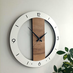 Op de afbeelding: Een moderne wandklok met een witte cirkelvormige lijst en een zwarte binnenring. De wijzerplaat heeft zwarte cijfers op 12, 3, 6 en 9 uur, met zwarte wijzers. Een verticaal houten paneel is gecentreerd op de wijzerplaat.