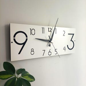May include: A rectangular white wall clock with black numerals and hands. The clock face displays the numbers 1 through 12 in a minimalist font. The clock hands are black and indicate the time. The clock is mounted on a white wall.