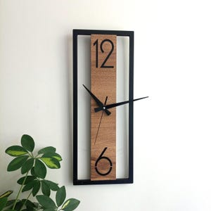 Könnte beinhalten: Eine rechteckige Wanduhr mit schwarzem Rahmen und Holzmitte. Das Zifferblatt zeigt die Zahlen 12 und 6 in Schwarz mit schwarzen Zeigern. Die Uhr ist an einer weißen Wand befestigt.
