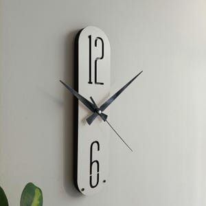 May include: A modern wall clock with a white face and black trim. The clock features black hands and the numbers 12 and 6 in a minimalist font. The clock is mounted on a light-colored wall.