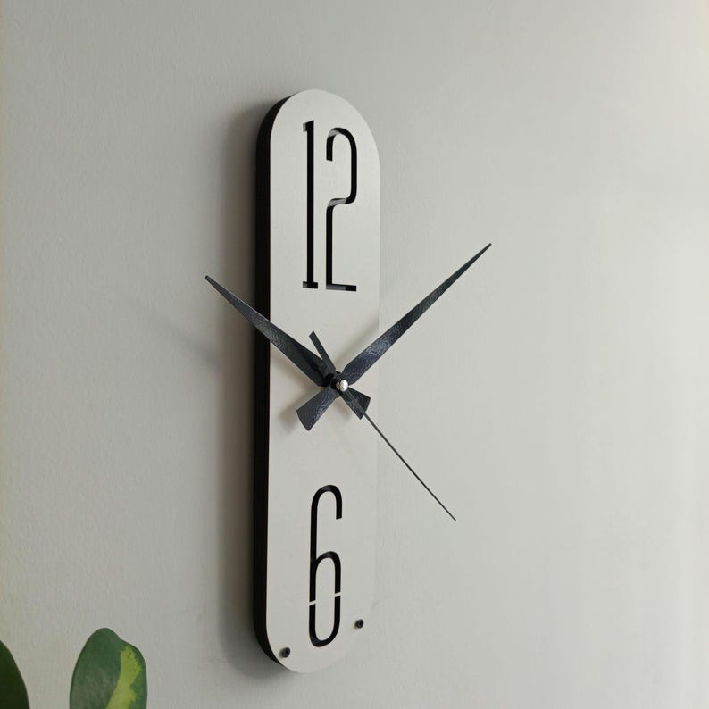 May include: A modern wall clock with a white face and black trim. The clock features black hands and the numbers 12 and 6 in a minimalist font. The clock is mounted on a light-colored wall.