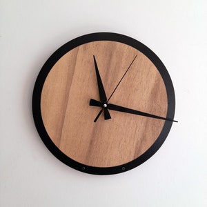 Puede incluir: Un reloj de pared redondo con una esfera de madera marrón claro, manecillas negras y un borde negro. El reloj está montado en una pared blanca. Las manecillas indican la hora.