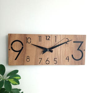 May include: A rectangular wooden wall clock with black numerals and hands. The clock face is a light brown colour with a wood grain pattern. The numbers 9 and 3 are large and bold, while the other numbers are smaller. The clock hands are black.