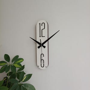 May include: A modern, minimalist wall clock with a white face and black hands and numerals. The clock has a long, oval shape and displays the numbers 12 and 6. The clock hands are black and pointed. The clock is mounted on a white wall.
