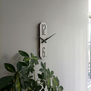 May include: A modern, minimalist wall clock with a white face and black hands and numerals. The clock features the numbers 12 and 6, and is mounted on a light gray wall. The clock's dimensions are not visible in the image.