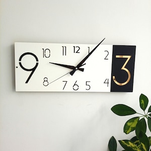 May include: A rectangular wall clock with a white face and black numerals. The clock has a black and gold accent on the right side with the number 3. The clock hands are black.