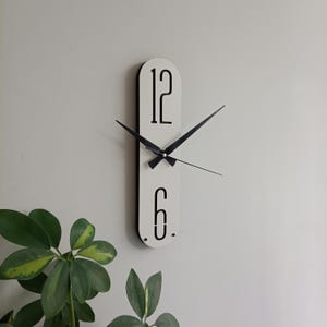 May include: A modern, minimalist wall clock. The clock has a white face with black numerals at the top and bottom, and black hands. The clock's shape is a long, rounded rectangle. The clock is mounted on a light gray wall.