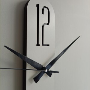 May include: A modern wall clock with a white face and black trim. The clock features black hands and the number 12 in a bold, minimalist font. The hands are positioned to show the time.
