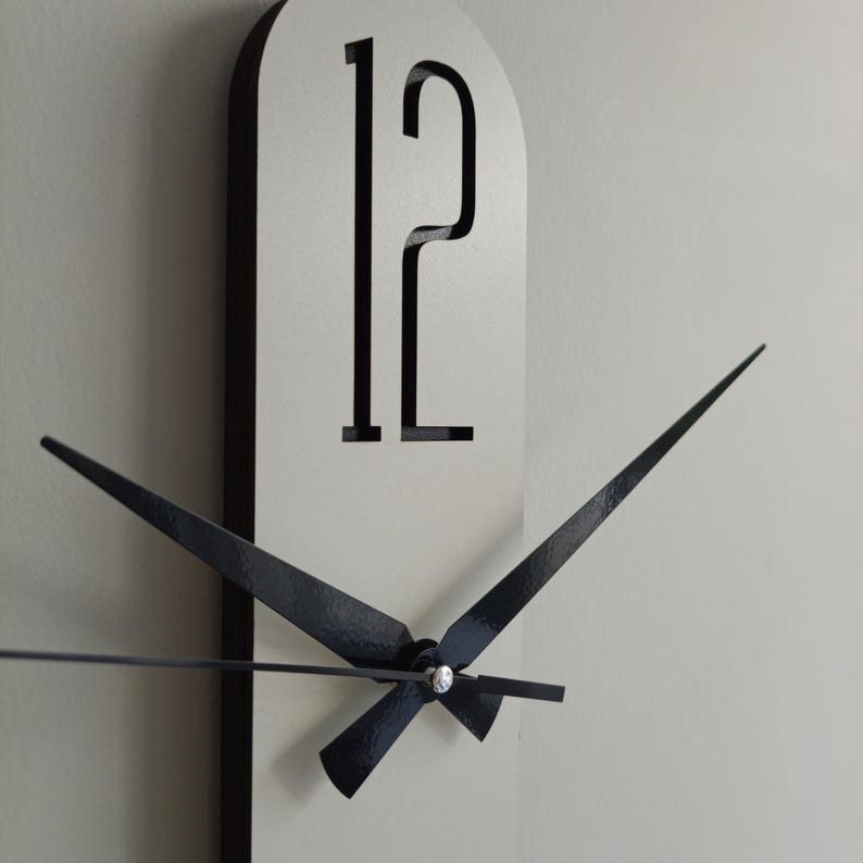 May include: A modern wall clock with a white face and black trim. The clock features black hands and the number 12 in a bold, minimalist font. The hands are positioned to show the time.