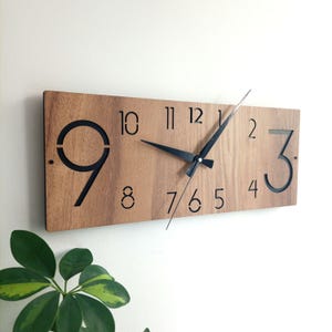 May include: A rectangular wooden wall clock with a light brown finish. The clock features large black numerals and black hands. The numbers 9 and 3 are open, while the other numbers are solid. The clock is mounted on a white wall.