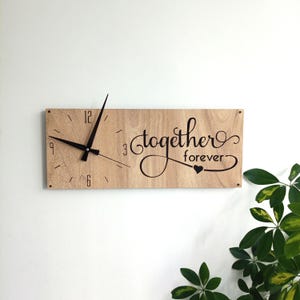 May include: A rectangular wooden wall clock with black hands and numerals. The clock face features the words "together forever" in a cursive font, with a heart symbol. The clock is mounted on a white wall.