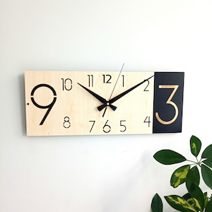 May include: A rectangular wall clock with a light wood face and black and gold accents. The clock features black hands, a black and gold number 3, and black numerals. The clock is mounted on a white wall.