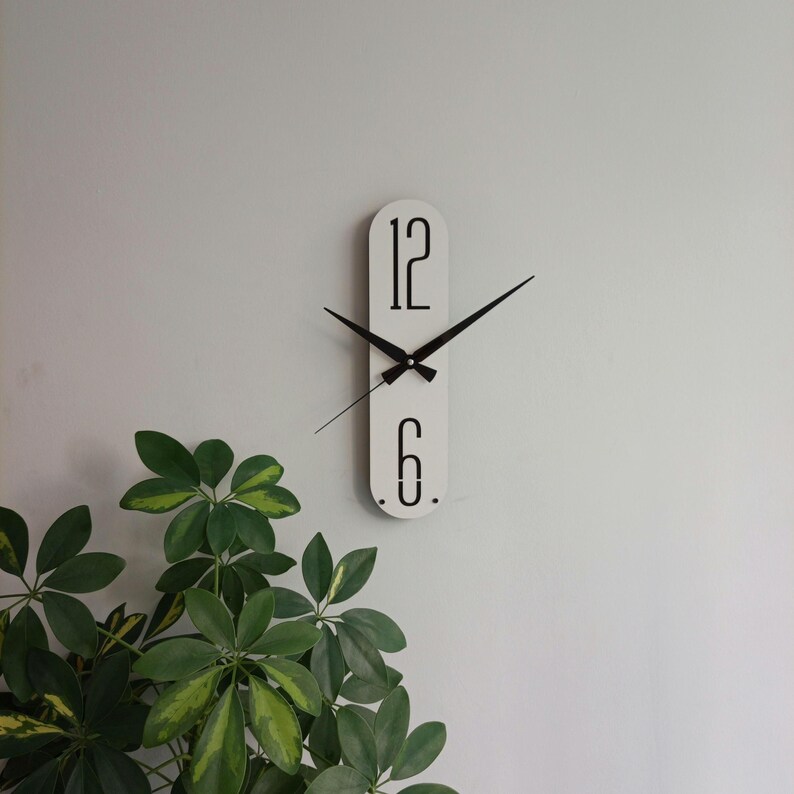 May include: A modern, minimalist wall clock. The clock has a white, elongated oval face with black hands and the numbers 12 and 6. The clock is mounted on a light gray wall, with green leafy plants in the foreground.
