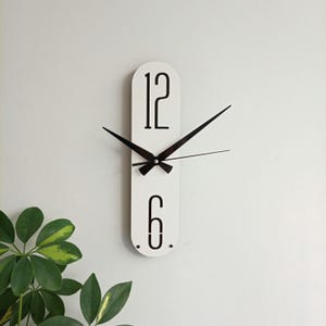 May include: White, elongated wall clock with black numerals and hands. The clock features the numbers 12 and 6, with a minimalist design. The clock's hands are black, and the background is a plain white wall. The clock is a decorative item.