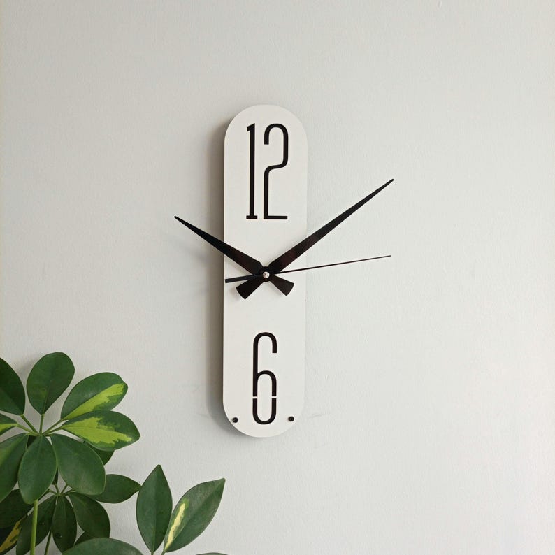 May include: White, elongated wall clock with black numerals and hands. The clock features the numbers 12 and 6, with a minimalist design. The clock's hands are black, and the background is a plain white wall. The clock is a decorative item.