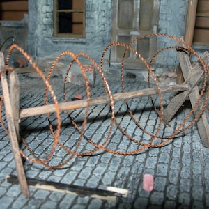 May include: A rusty barbed wire fence is coiled and secured to a wooden post. The fence is made of multiple strands of wire twisted together. The post is made of rough-hewn wood and is weathered and worn. The fence is set against a backdrop of a brick wall and a cobblestone street.