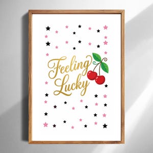 May include: A framed print with a white background, featuring the words "Feeling Lucky" in gold script. The image includes red cherries with green leaves and scattered pink and black stars.
