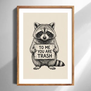 May include: A framed art print featuring a detailed illustration of a raccoon holding a sign. The sign reads "TO ME YOU ARE TRASH" in a bold, readable font. The artwork is in black and white.