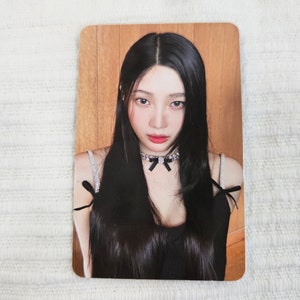 May include: A rectangular photo card featuring a woman with long black hair and red lipstick. She wears a black top with rhinestone straps and a black bow choker. The background is a light brown wood panel.